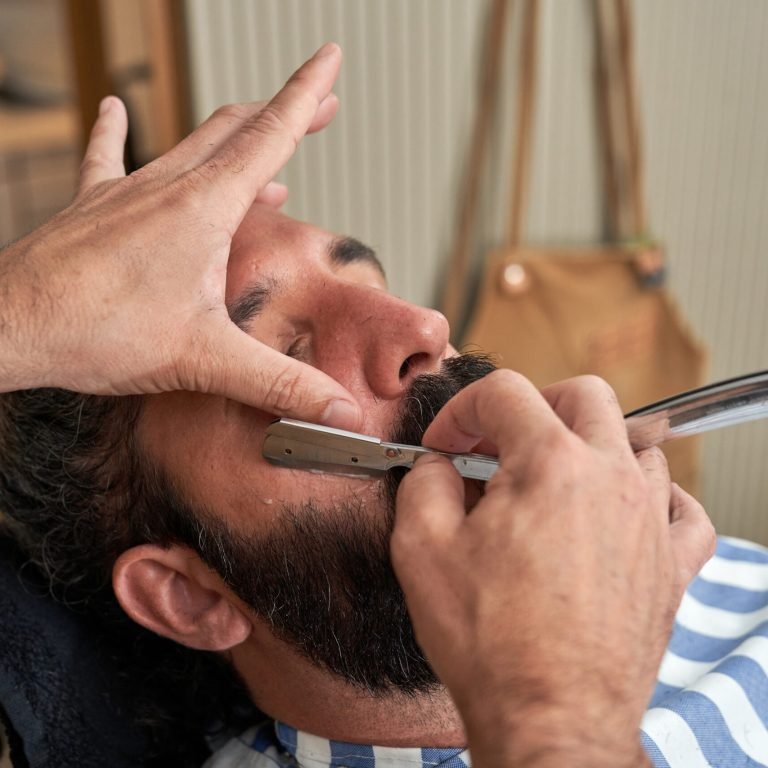 Crop barber with tattoo cutting beard of client with straight blade while working in barbershop